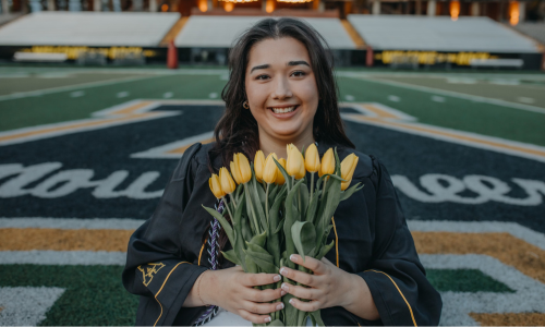  Melissa Castaneda in her cap and gown holding yellow flowers on the football field at App State.